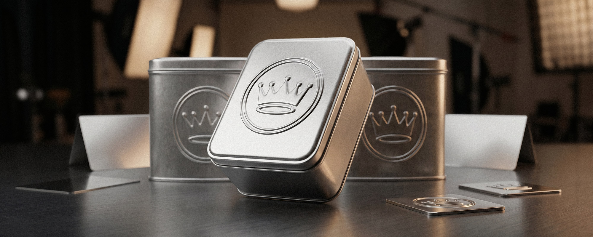 Mehrere Metall-Dosen mit geprägtem Kronenmotiv auf dunkler, reflektierender Oberfläche; eine Dose im Vordergrund, weitere im Hintergrund, dazu kleine Metallplättchen mit Emblem.