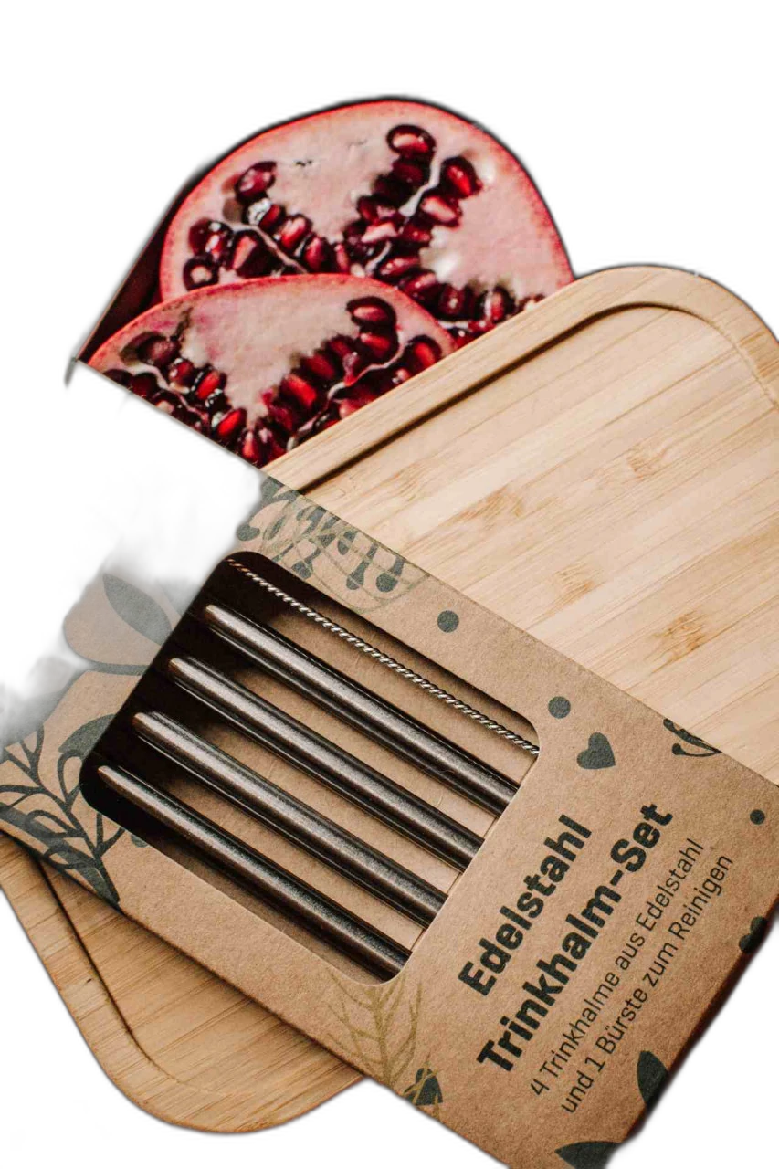Nachhaltiges Edelstahl Trinkhalm-Set in Kartonverpackung auf einem Bambusdeckel, im Hintergrund eine geöffnete Metalldose mit frischen Granatapfelhälften und Blaubeeren auf Holztisch.