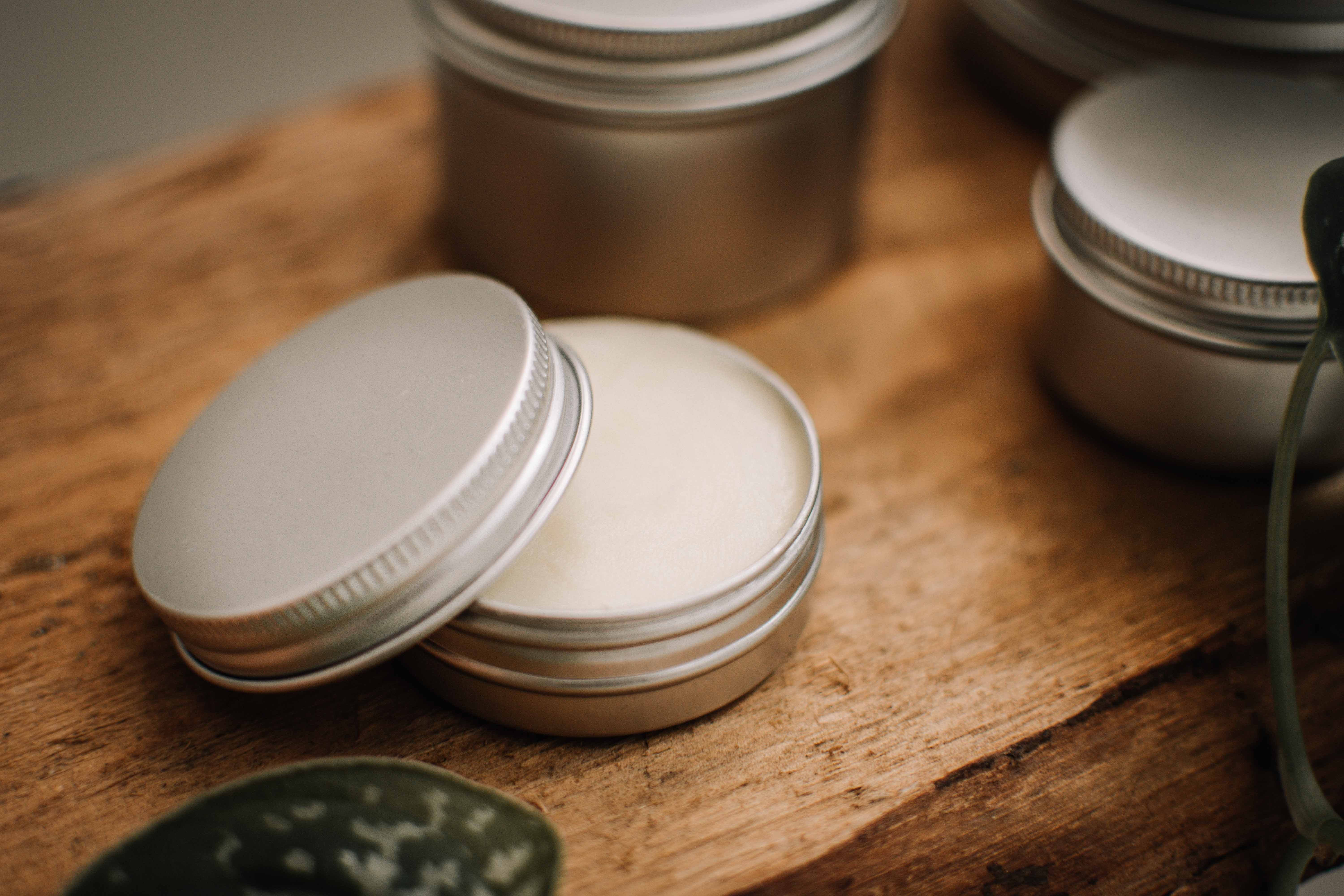 Geöffnete silberne Aluminiumdose mit 30 ml Inhalt, gefüllt mit einer weißen Creme oder Salbe, stehend auf einer rustikalen Holzoberfläche, umgeben von weiteren geschlossenen Dosen und einer Pflanze im Vordergrund.