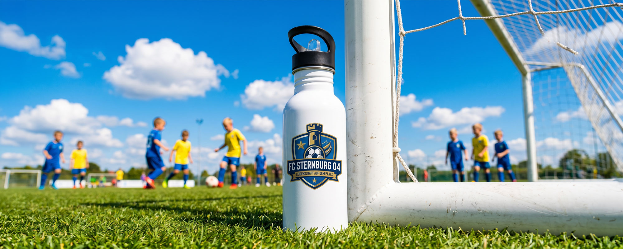 Weiße Edelstahl-Trinkflasche mit Vereinslogo „FC Sternburg 04“ am Fußballtor, vor spielenden Kindern auf dem Fußballplatz.