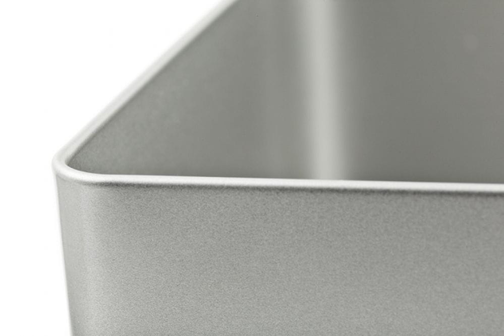 Geöffnete quadratische Blechdose aus silberfarbenem Metall mit abgenommenem Stülpdeckel, Detailaufnahme einer Ecke, auf weißem Hintergrund fotografiert.