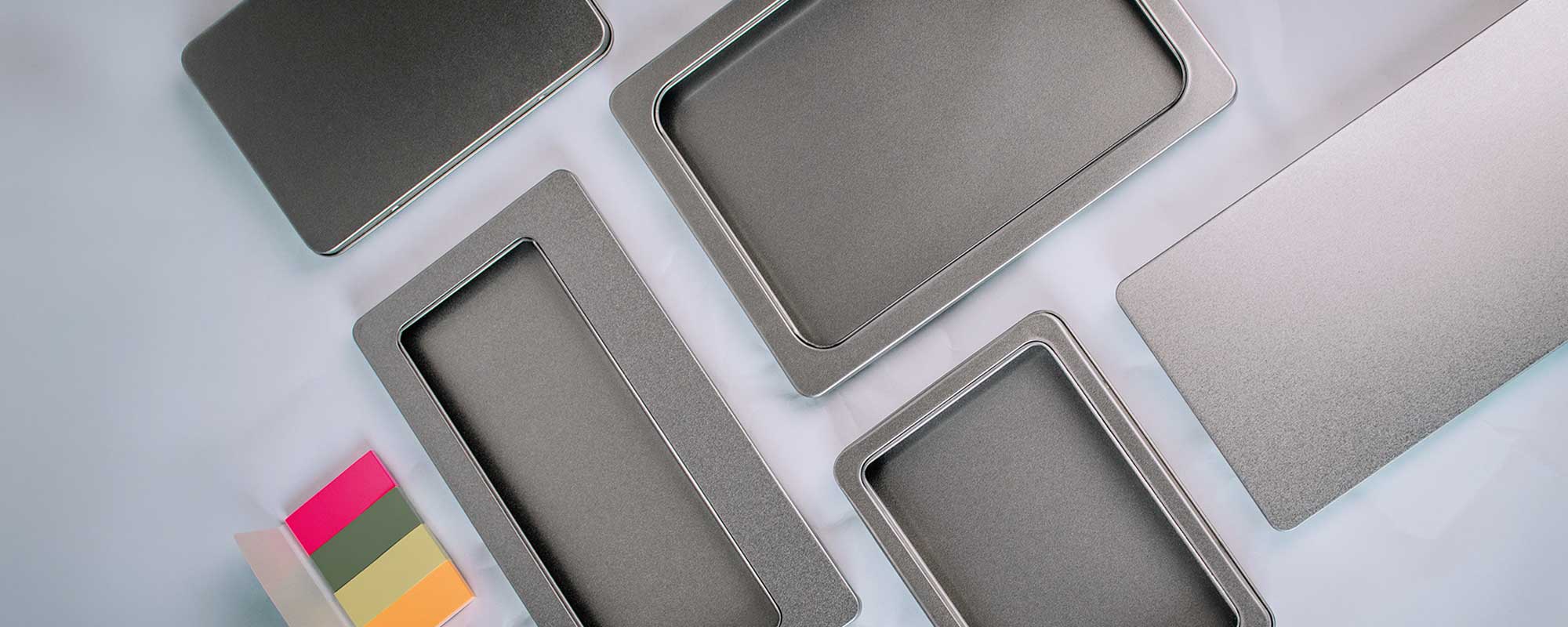 Sammlung rechteckiger Metalldosen mit Sichtfenstern auf grauer Oberfläche; moderne Produktaufnahme mit Farbmuster-Karten als Akzent.