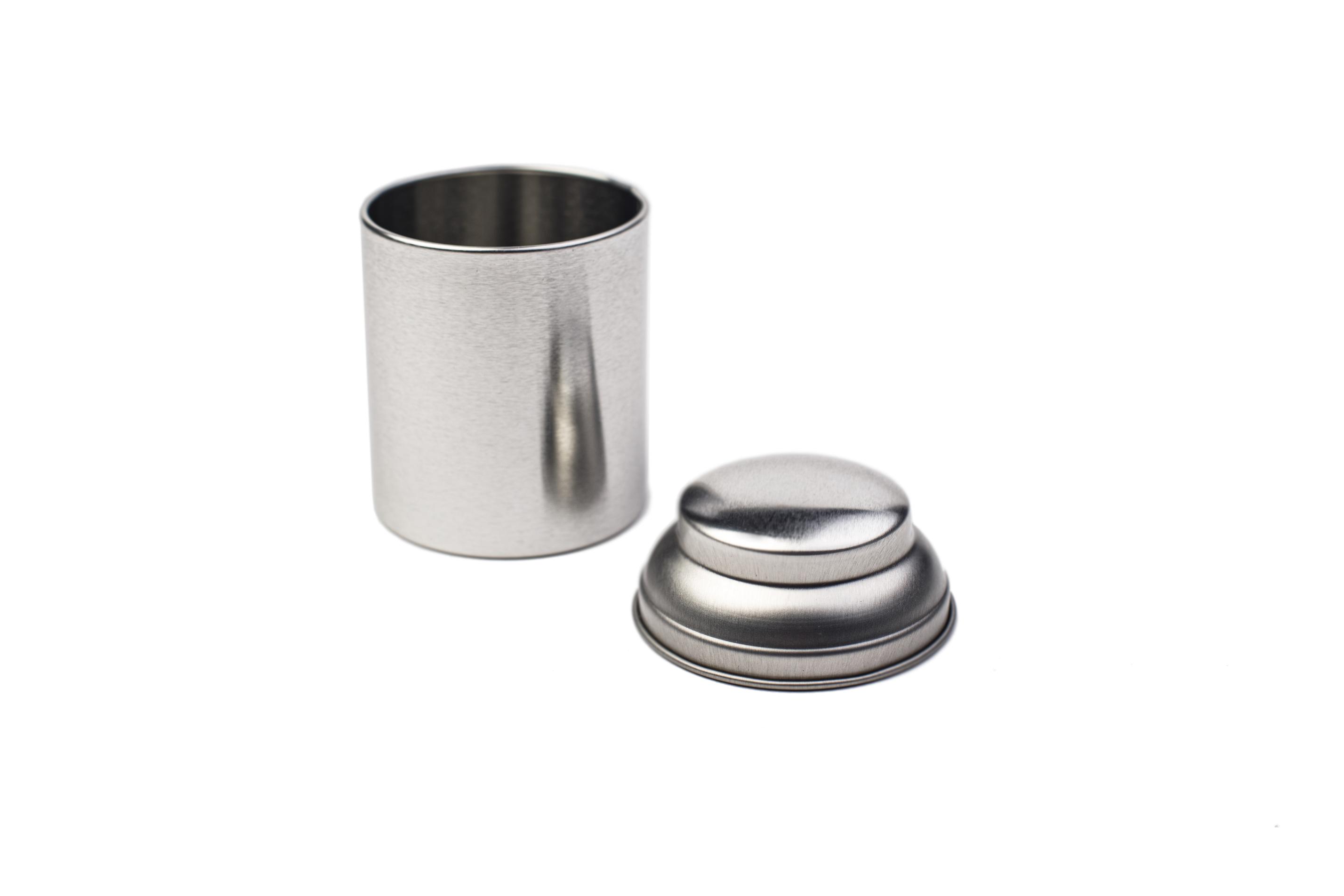 Geöffnete kleine runde Teedose aus silberfarbenem Metall, mit abgenommenem Deckel, auf weißem Hintergrund fotografiert.