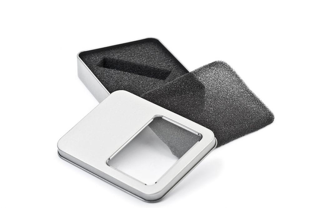 Geöffnete rechteckige silberfarbene Metalldose mit transparentem Sichtfenster im Deckel, schwarzer Schaumstoffeinsatz für USB-Stick, auf weißem Hintergrund fotografiert.