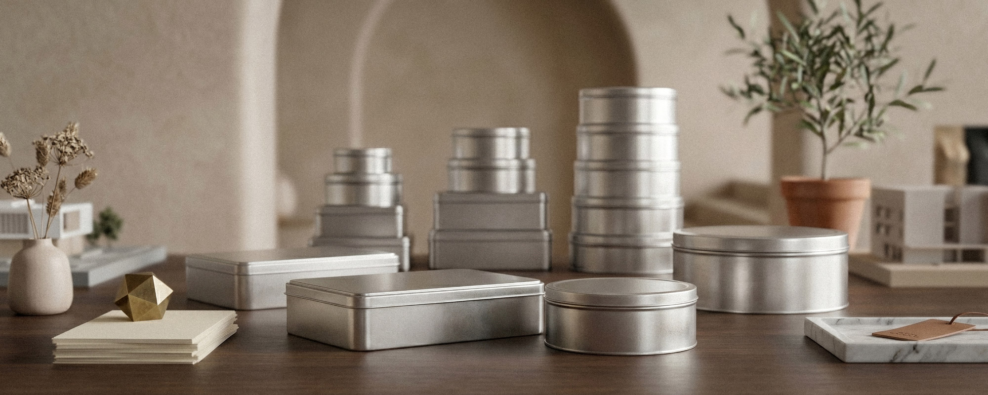 Sammlung silberner Metalldosen in verschiedenen Formen und Größen auf Holztisch; stilvolle Produktaufnahme mit Vase, Karten, Golddeko und Olivenbaum im Hintergrund.