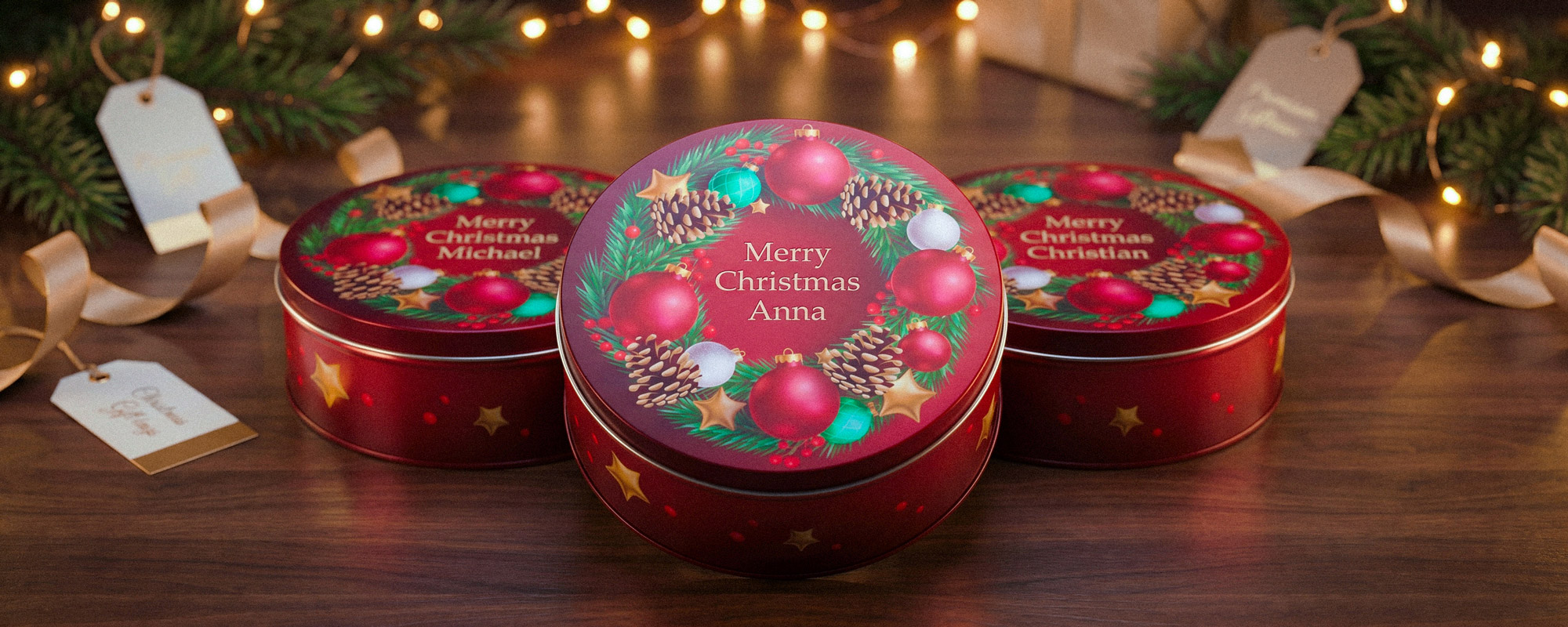 Drei personalisierte Weihnachtsdosen in Rot mit Kranzmotiv, goldenen Sternen und „Merry Christmas“-Aufdruck; festliche Produktaufnahme mit Tannenzweigen und Lichterkette.