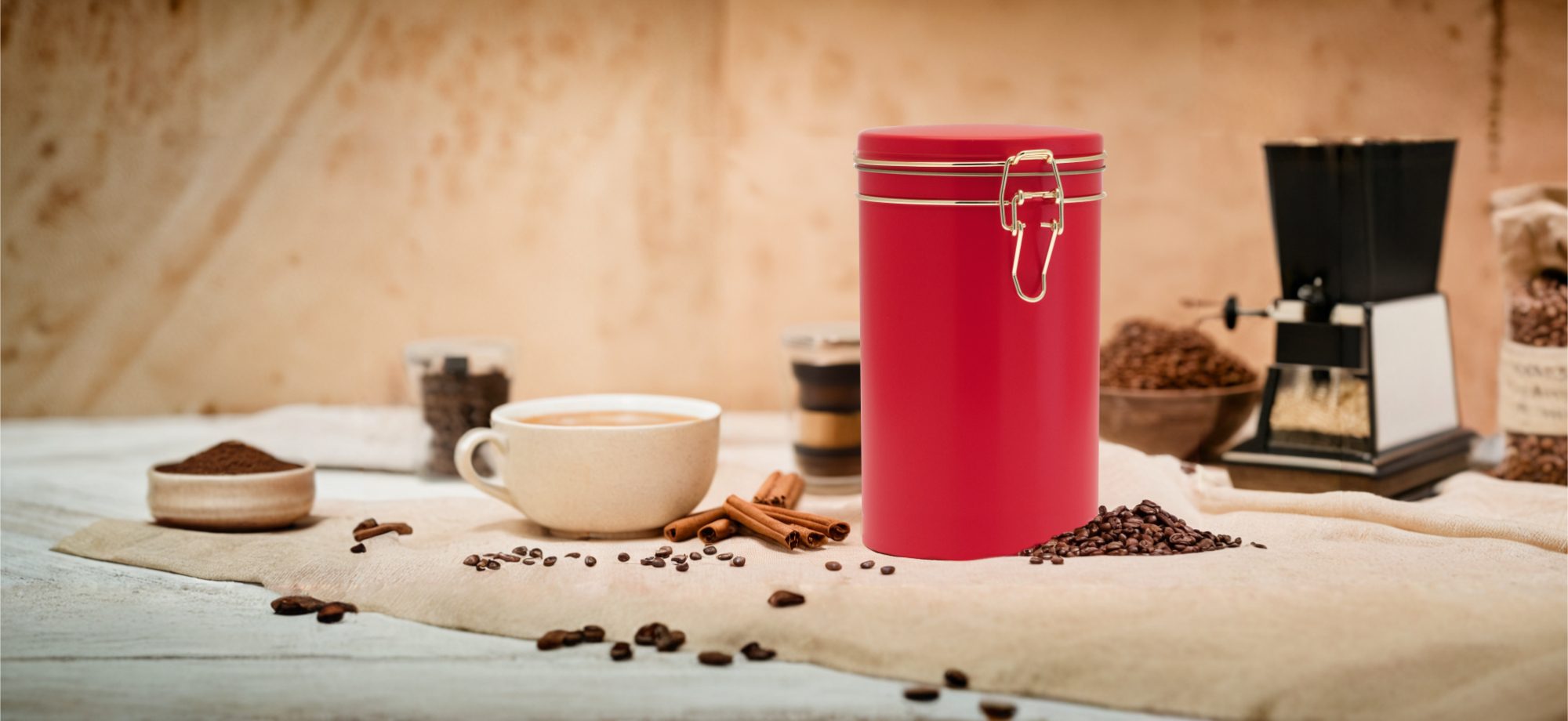 Rote Kaffeedose mit Metallverschluss auf Stoffunterlage, umgeben von Kaffeebohnen, Zimtstangen, einer Tasse Kaffee, Kaffeepulver und einer manuellen Kaffeemühle. Warme, rustikale Produktaufnahme.