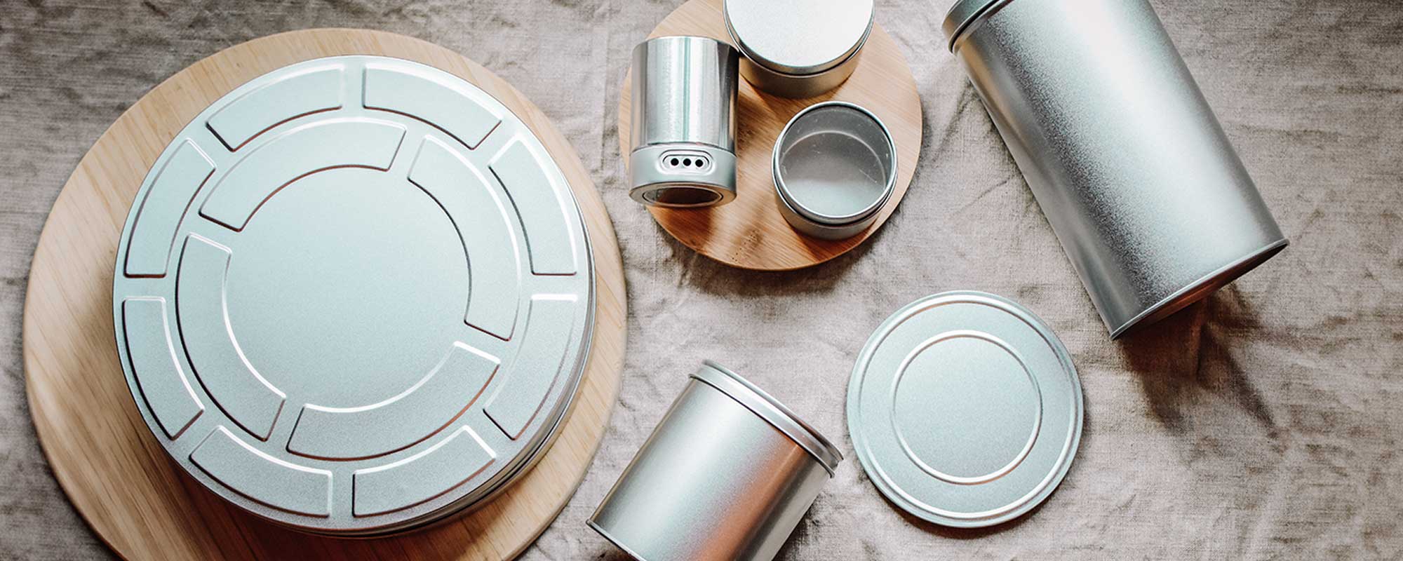 Verschiedene silberne Metalldosen und Deckel auf Leinenstoff und Holzunterlagen; stilvolle Produktaufnahme mit Fokus auf Formen, Oberflächen und Materialqualität.