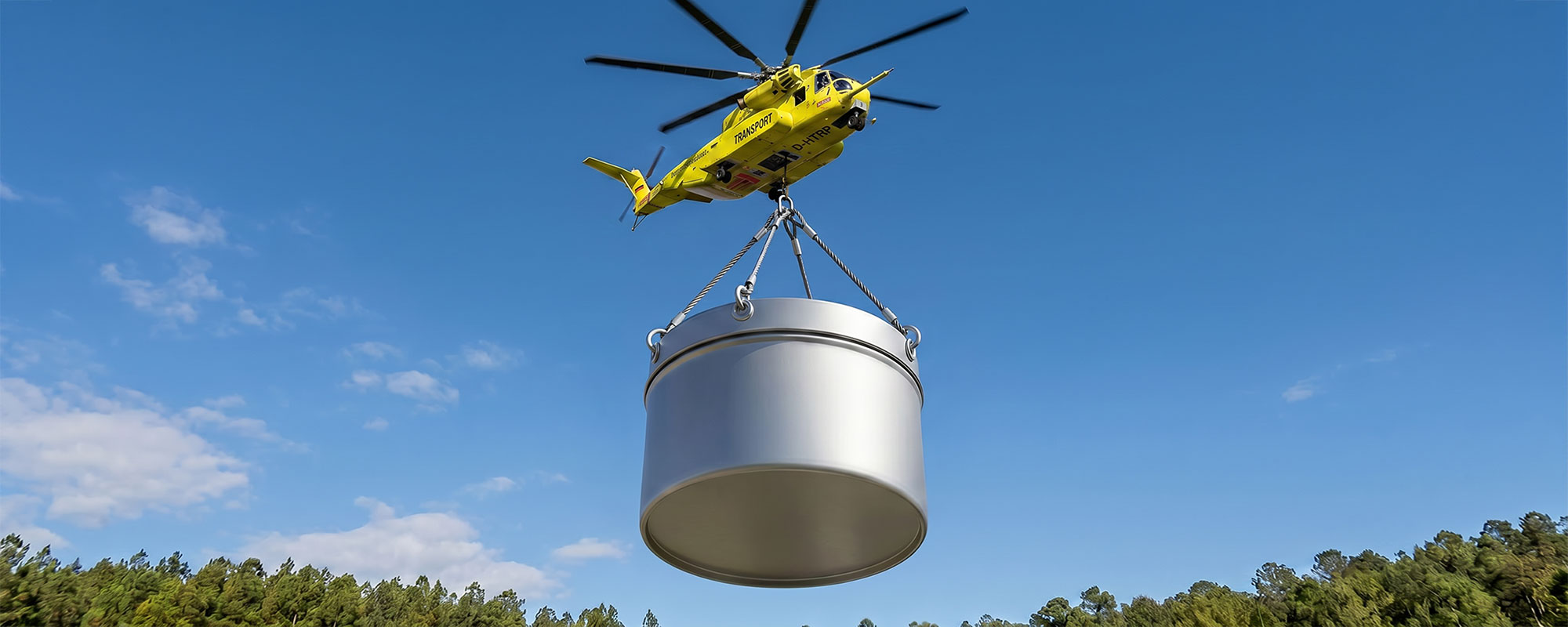 Transporthubschrauber hebt eine große silberne Metalldose per Seiltransport über einer Waldlandschaft; dynamische Aufnahme vor blauem Himmel.